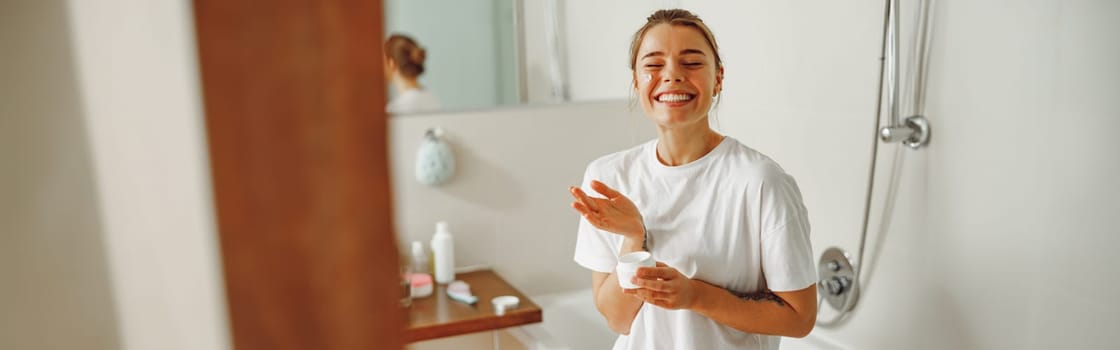 Smiling woman holding jar with skincare cream in hands. Home beauty routine