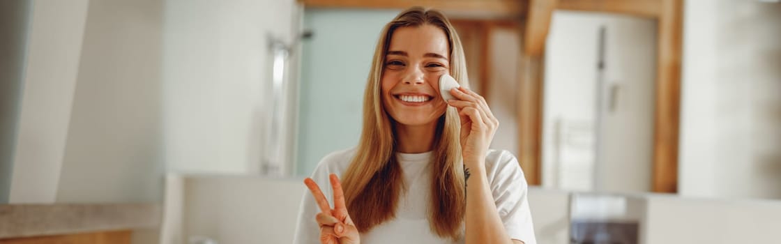 Young woman removing makeup with cotton pad standing in bathroom. Home beauty routine