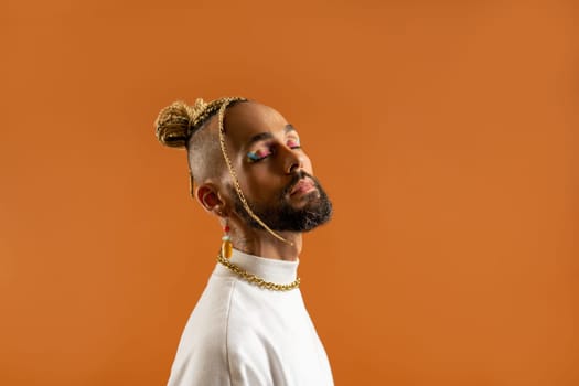 Confident black gay man with bright makeup standing isolated on orange background, dressed white. Exudes sense of pride and individuality. Diversity power of personal style. Closed eyes