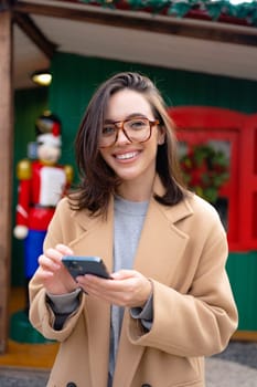 Woman in glasses using mobile phone at day in the city. Happy woman using smartphone dressed stylish trench coat looking at camera. Vertical photo portrait