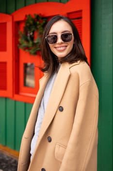 People, season and Christmas concept portrait of happy smiling middle age woman outdoors dressed stylish cream trench coat with Christmas festive decoration on background. Vertical photo, portrait