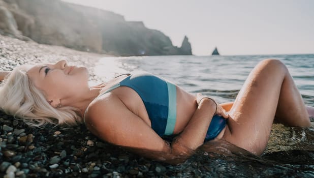 A woman in a blue bikini lies on a rocky beach, basking in the sun. She has blonde hair, and the water is just below her.