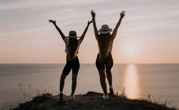 Two women in hats stand with arms raised on a cliff overlooking the ocean at sunset. The setting sun is reflected on the water below.