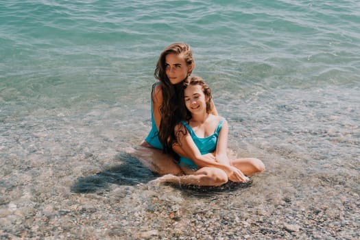 Two women in blue bathing suits are relaxing in shallow, clear ocean water.