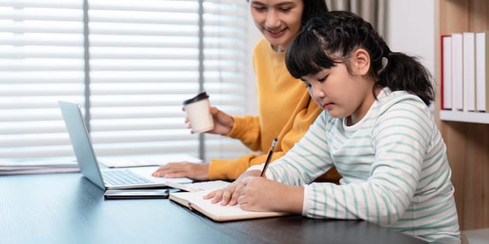 A mother and daughter spending quality time together at home, with the mother working on a laptop and the daughter studying.