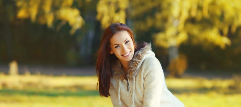 Portrait of happy young woman smiles in sweater looking at camera in sunny autumn park with yellow leaves