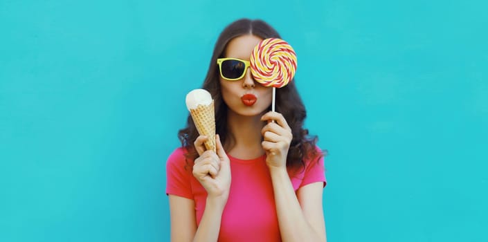 Summer portrait of happy young woman holding ice cream cone and lollipop blowing a kiss on blue background
