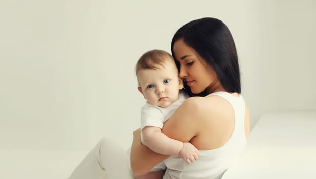 Happy young mother holding her cute baby sitting together in white room at home