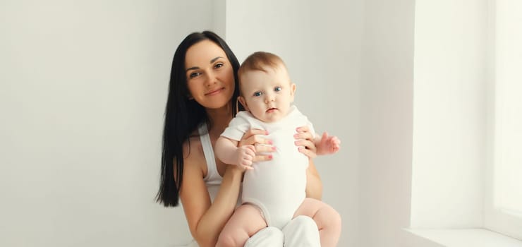 Happy smiling young mother playing with cute baby sitting together in white room at home