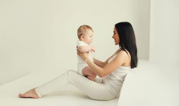 Happy smiling young mother playing with cute baby sitting together in white room at home