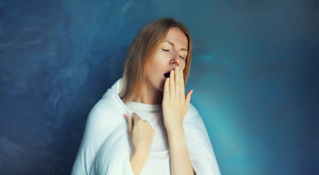 Sleepy lazy young woman yawns waking up after sleeping wrapped in white soft comfortable blanket in morning standing on blue background at home
