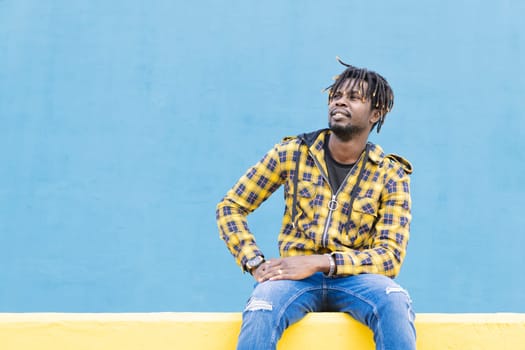 smiling black man sitting on a yellow wall with a blue wall as a background, lifestyle concept, copy space for text