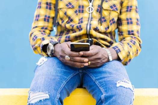 hands of an unrecognizable black man writing a message on his mobile phone sitting on a yellow wall with a blue wall in the background, technology and lifestyle concept, copy space for text