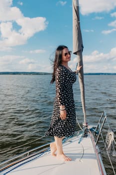 A woman in a patterned dress and sunglasses stands on a sailing boat under a clear sky