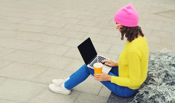 Modern young woman working on a laptop, freelancer girl looking at computer in colorful clothes, cup of coffee drink or juice sitting on a city street