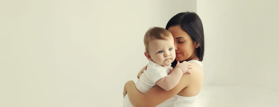 Happy young mother holding her cute baby sitting together in white room at home