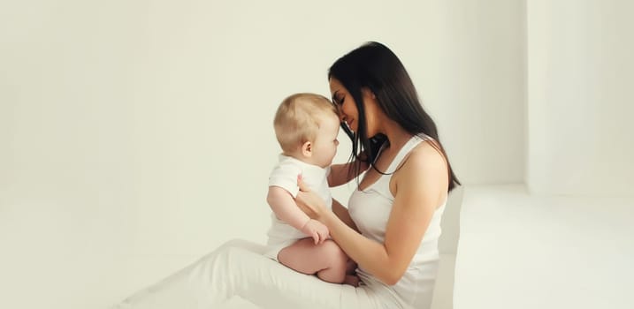 Happy young mother holding her cute baby sitting together in white room at home