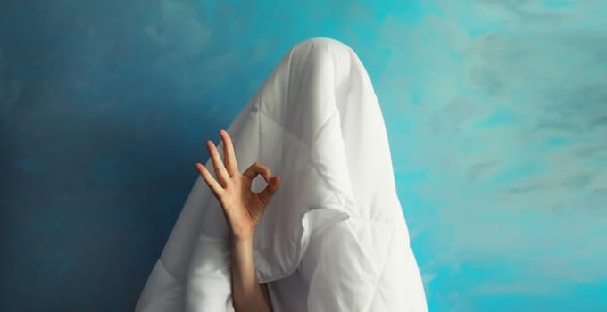 Cheerful lazy woman waking up after sleeping covered head with white blanket showing ok gesture her fingers on blue background