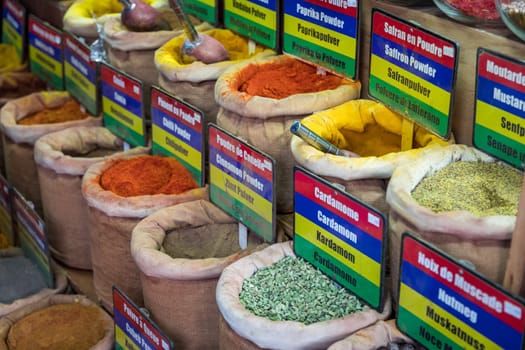 A colorful display of spices and herbs, including cumin, coriander, and ginger. The spices are arranged in small bags and baskets, and there are signs next to each one indicating their names and uses
