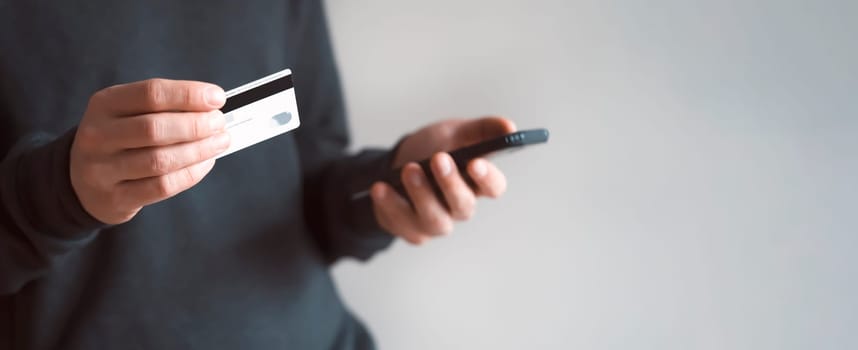 Male hands are holding a mobile phone and a bank card, a young man in a dark casual sweater pays for purchases, makes online banking using an application, close-up view.