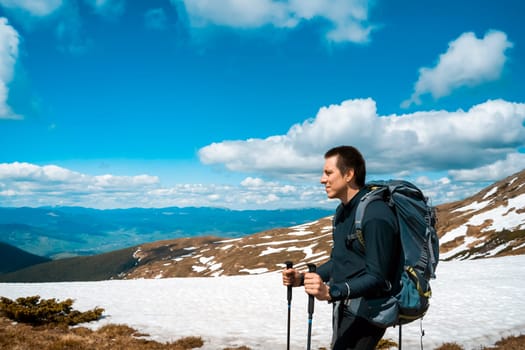 A young man, while hiking in the mountains in winter, looks ahead and admires the stunning mountain landscape. A hiker with tourist equipment climbs to the top of a mountain in a national park.