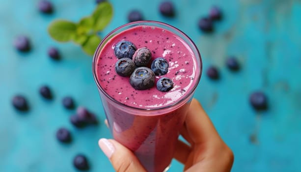 Woman is holding a glass of blueberry smoothie decorated with fresh blueberries on a blue background