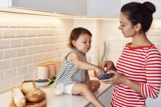 Little baby girl with her mother eating fresh blueberries in white kitchen