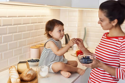 mother gives to the little baby girl fresh blueberries in white kitchen