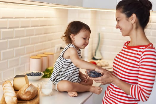 Little baby girl with her mother eating fresh blueberries in white kitchen