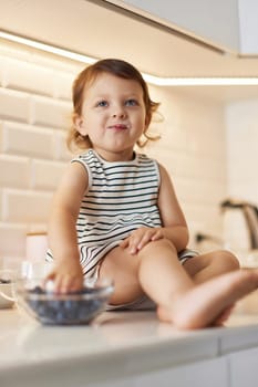 Little baby girl eating fresh blueberries from glass bowl in white kitchen