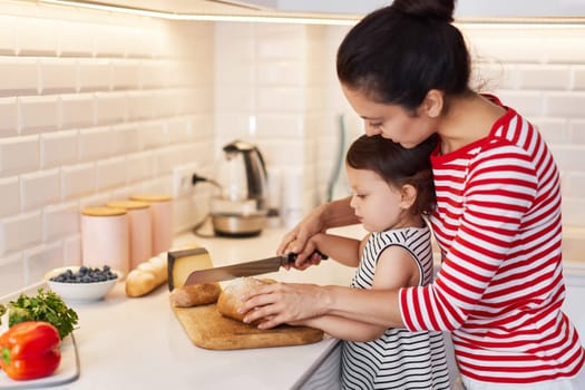 mother helps her cute little baby girl with knife for education in modern white kitchen