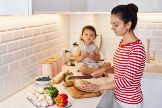 mother and cute little child daughter preparing breakfast in modern white kitchen. mom cutting bread on table.