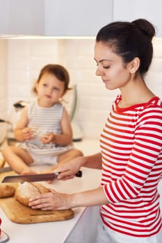 mother and cute little child daughter preparing breakfast in modern white kitchen. mom cutting bread on table.