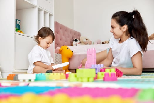 cute baby girl and her loving mother playing with toy bricks in the children room, early development