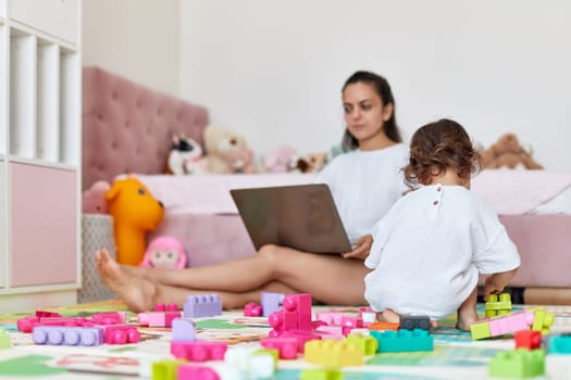 little baby girl playing with toys blocks and working mother sitting on floor with laptop in room