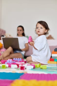 cute little baby girl playing with toys and mom sitting on floor with laptop in room