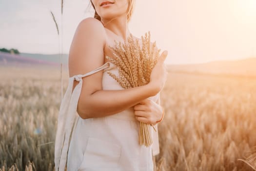 A woman is holding a bunch of wheat in her arms. The wheat is dry and brown, and the woman is wearing a white dress. The scene is set in a field, and the woman is posing for a photo