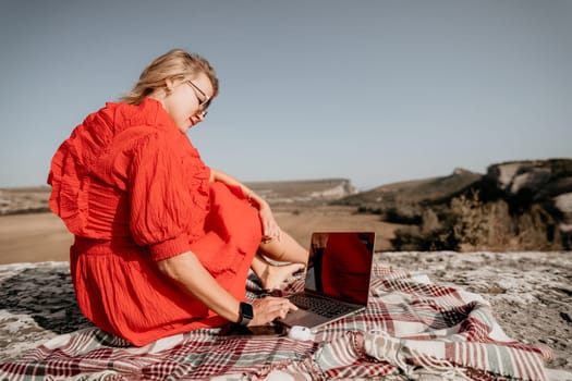A woman in a red dress sits on a blanket, working on a laptop, against the backdrop of a mountain landscape.