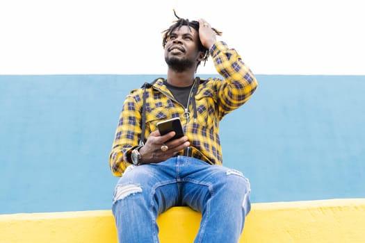 smiling black man sitting on a yellow wall with a smart phone at his hand, concept of technology and lifestyle, copy space for text