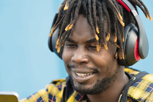 portrait of a young african man with headphones consulting his smart phone with a blue wall in the background, technology and lifestyle concept, copy space for text