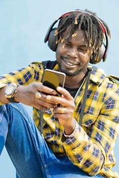 vertical portrait of a young black man with a headphones writing on his smart phone with a blue wall in the background, technology and lifestyle concept, copy space for text