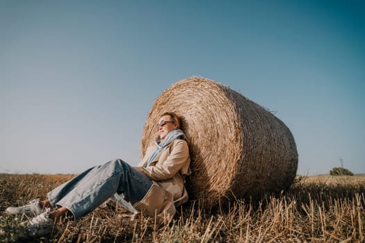 A young woman wearing sunglasses and a beige coat leans back against a hay bale in a field.