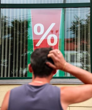 A man is standing outside a store, scratching his head as he observes a large percentage sign displayed in the window. His posture suggests he might be puzzled or considering the implications of the advertised discount, sale, or promotion.