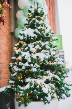 Closeup of festively decorated outdoor Christmas tree with bright balls on winter city background. Garland lights and winter snow - holidays xmas festive