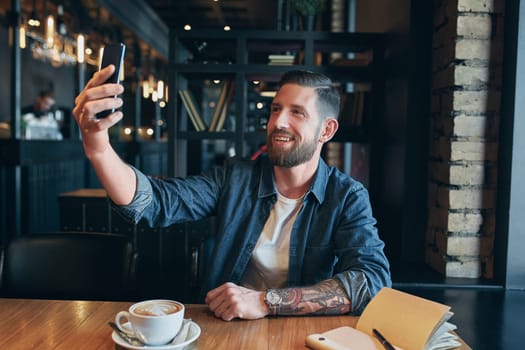 Man looking into the camera while making a selfie. Young bearded man, dressed in a denim shirt, sitting at table in cafe and use smartphone
