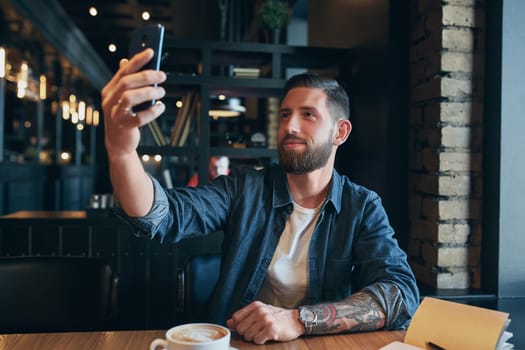 Man looking into the camera while making a selfie. Young bearded man, dressed in a denim shirt, sitting at table in cafe and use smartphone