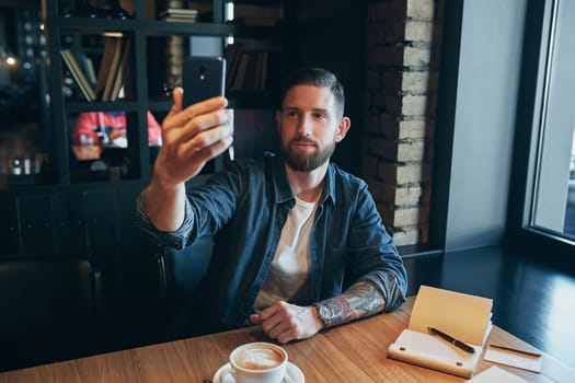 Man looking into the camera while making a selfie. Young bearded man, dressed in a denim shirt, sitting at table in cafe and use smartphone