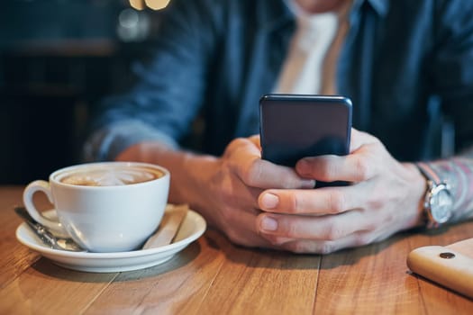 Man hands in denim shirt slide with finger on screen his smart phone, near cup with coffee on wooden table in cafe shop during his coffee break