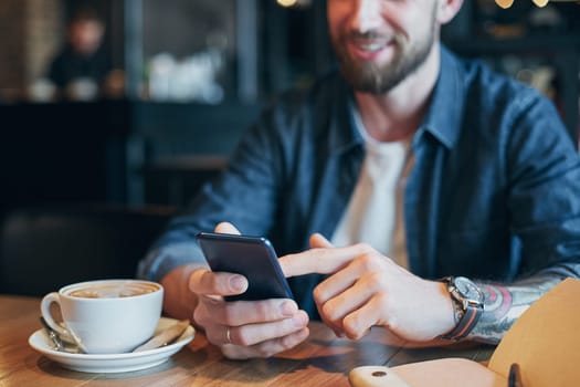 Man hands in denim shirt slide with finger on screen his smart phone, near cup with coffee on wooden table in cafe shop during his coffee break