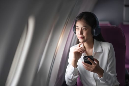 Asian woman sitting and using a smartphone in an airplane Listening to music next to the airplane seat window travel and technology concept.